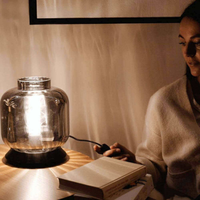 Frau, die an einem Tisch mit einer dekorativen Lampe und einem Buch sitzt, in gemütlicher Atmosphäre.