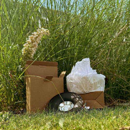 Braune Kartons und Verpackungsmaterial in der Wiese, umgeben von hohem Gras und Pflanzen.