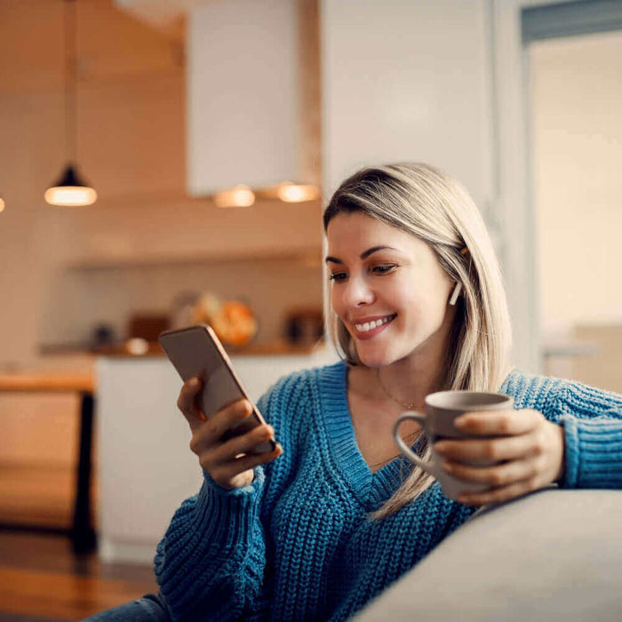 Junge Frau sitzt auf dem Sofa, schaut lächelnd auf ihr Smartphone und hält eine Tasse in der Hand.