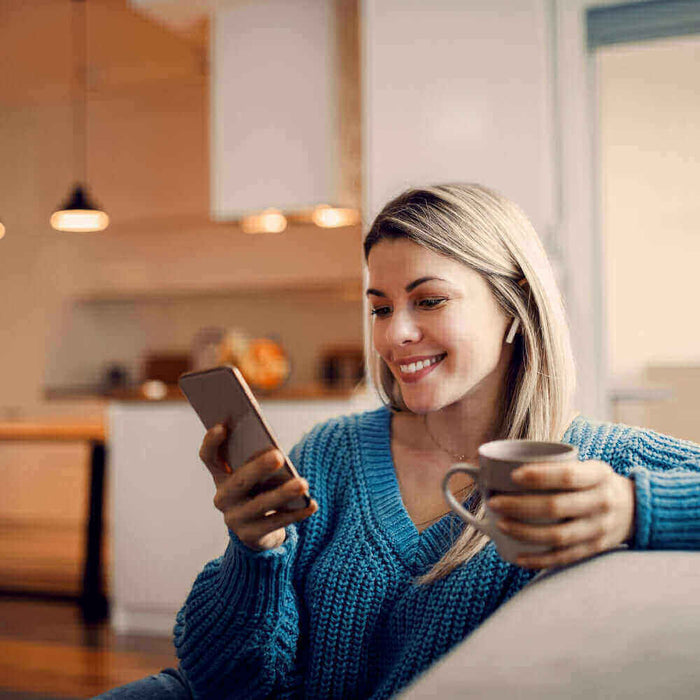 Junge Frau sitzt auf dem Sofa, schaut lächelnd auf ihr Smartphone und hält eine Tasse in der Hand.