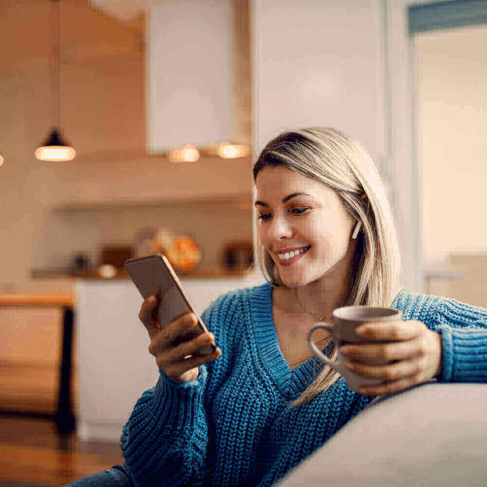 Junge Frau sitzt auf dem Sofa, schaut lächelnd auf ihr Smartphone und hält eine Tasse in der Hand.