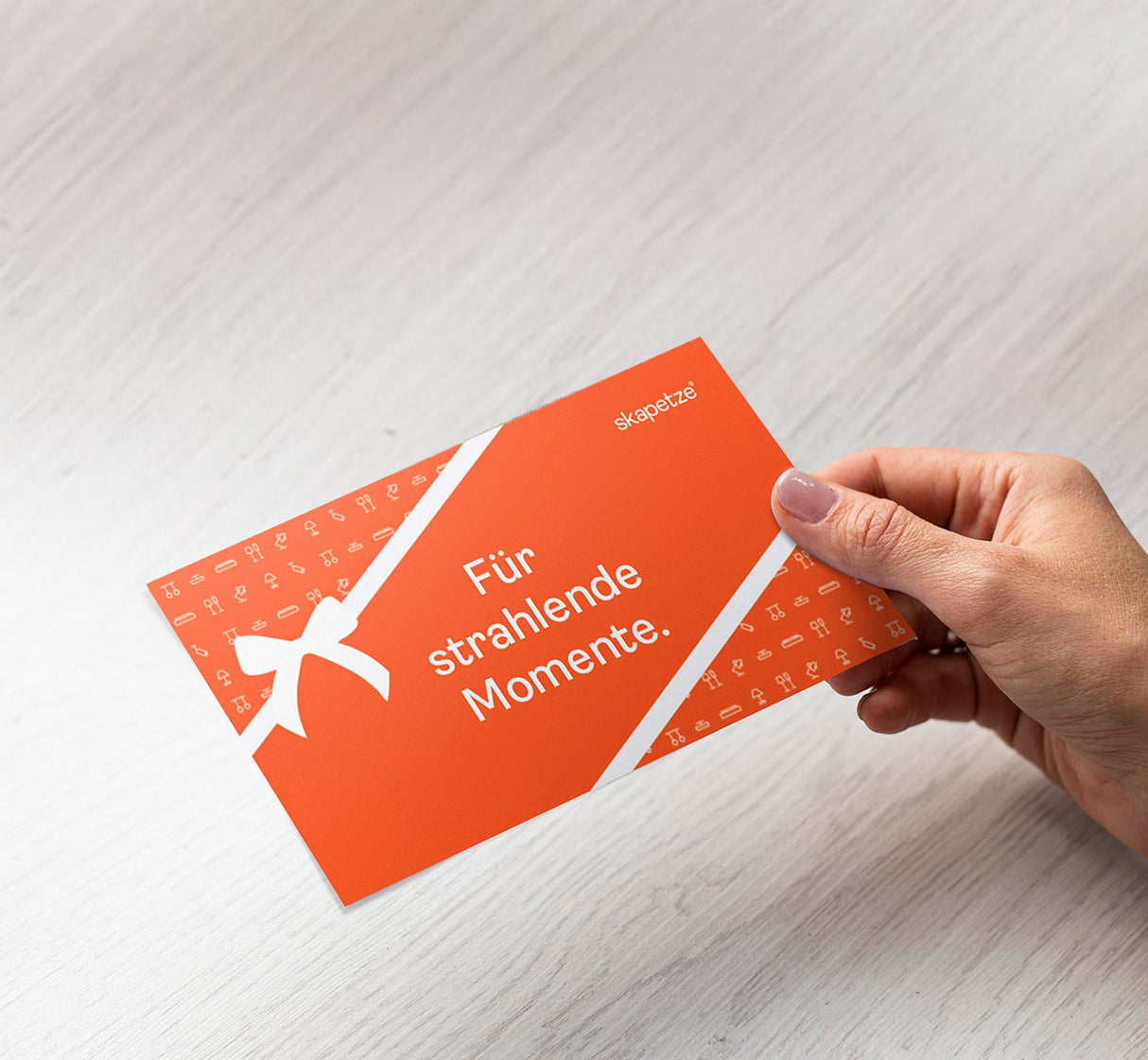 Hand hält den skapetze® Leuchten-Gutschein mit dem Text "Für strahlende Momente" auf orangefarbenem Hintergrund.