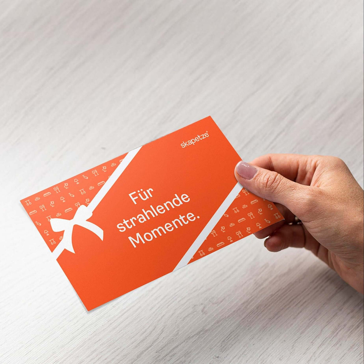 Hand hält den skapetze® Leuchten-Gutschein mit dem Text "Für strahlende Momente" auf orangefarbenem Hintergrund.