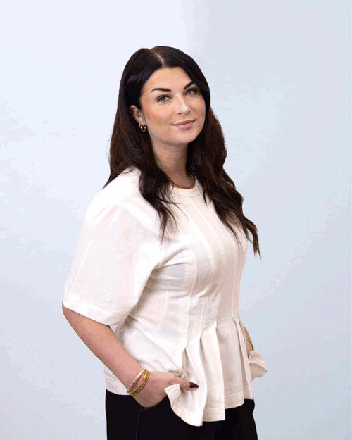 Porträt einer Frau mit langen, dunklen Haaren, die ein helles Oberteil trägt, vor einem neutralen Hintergrund.