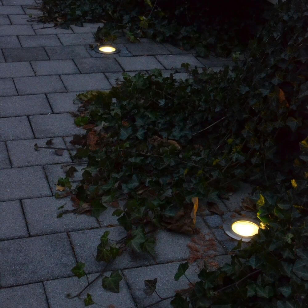 Runde Edelstahl Bodeneinbaustrahler im Gartenweg, umgeben von Efeu, beleuchten den schattenhaften Bereich.