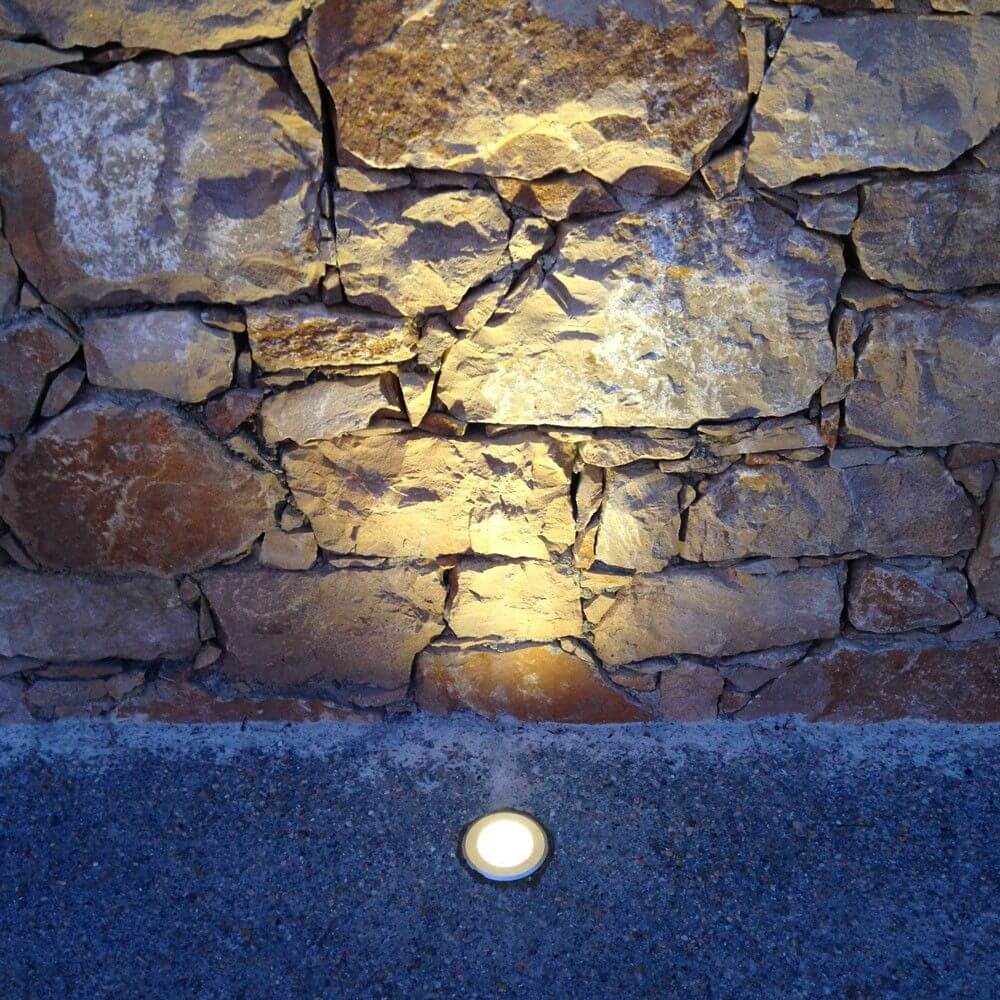 Runder Level Bodeneinbaustrahler aus Edelstahl, strahlend auf Steinwand, IP67 Schutz.