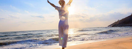 Frau mit ausgebreiteten Armen am Strand, Sonnenlicht strahlt hinter ihr, symbolisiert Freiheit und Lebensfreude.