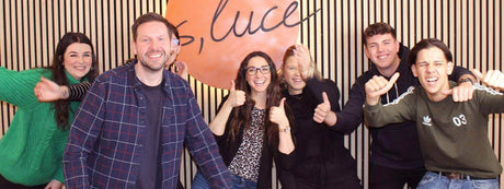 Gruppe von sechs Personen mit Daumen hoch und Lächeln, stehen vor einem orangefarbenen Hintergrund mit 'luce'-Schriftzug.