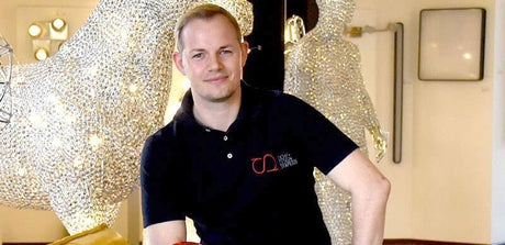 Guido Huber von skapetze® posiert in einem hellen Raum mit glänzenden Skulpturen im Hintergrund.