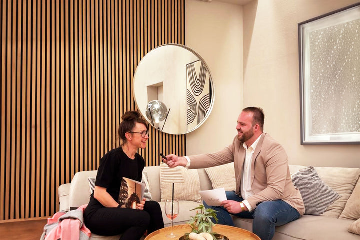 Anja Gödecke sitzt links auf einem Sofa und wird von einem skapetze®-Mitarbeiter mit einem Mikrofon interviewt, in einem modern eingerichteten Raum mit Holzlamellenwand und Spiegel.