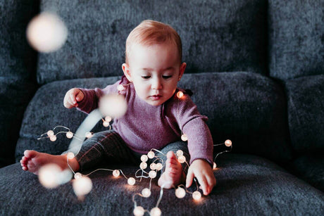 Kleines Kind sitzt auf Sofa und spielt mit Lichterketten in warmem Licht, schafft eine gemütliche Atmosphäre.