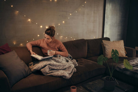 Gemütliche Atmosphäre mit warmem Licht. Frau liest auf der Couch mit einer Tasse in der Hand, umgeben von Kissen.