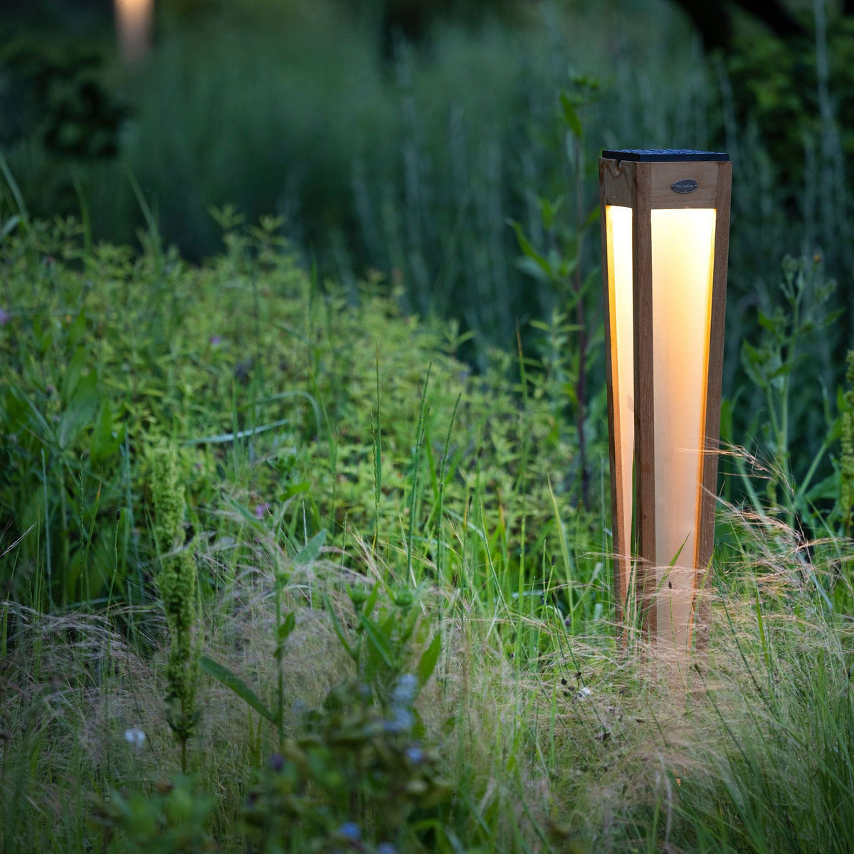 Antorcha solar Tecka de madera de teca, 120 cm de alto, color natural, iluminando el jardín al anochecer