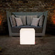 Moree Cube Outdoor LED Sitzwürfel vor einer gemütlichen Sofa-Ecke im Freien bei gedämpfter Beleuchtung.