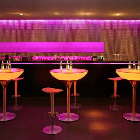 Stół LED Moree Lounge Table LED Pro 105 cm w nowoczesnym wnętrzu z wielokolorowym oświetleniem i stylowymi hokerami.
