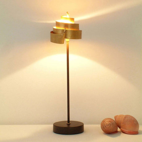 Tischleuchte Piccola Banderole Eisen Braun-Schwarz-Gold auf einem Tisch mit Muscheln und warmem Licht