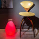 Akku LED-Dekolampe Vessel S in Rot neben einem Holzstuhl, modernes Design und stimmungsvolle Beleuchtung.