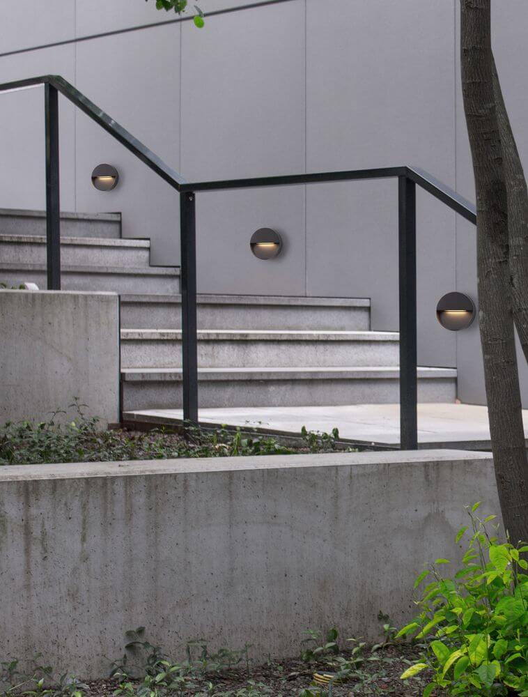 Arne LED Außenwandleuchte Ø 12cm IP54 an einer Treppe in moderner Umgebung mit Beton und Pflanzen.