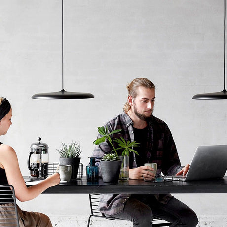 Sospensione Artist 25 LED Ø 25 cm dimmerabile nera sopra un moderno tavolo da pranzo con piante e laptop.