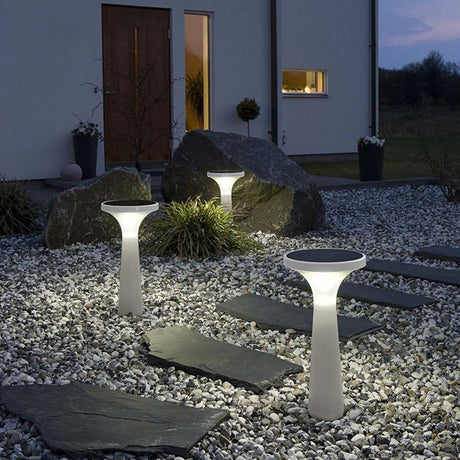 Luz decorativa LED solar Assisi Aton 45cm blanca en el jardín con camino de piedra y iluminación nocturna.