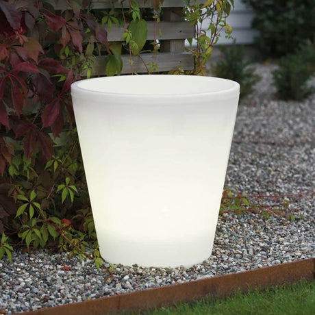 Assisi LED planter glows softly in the garden among plants and gravel.