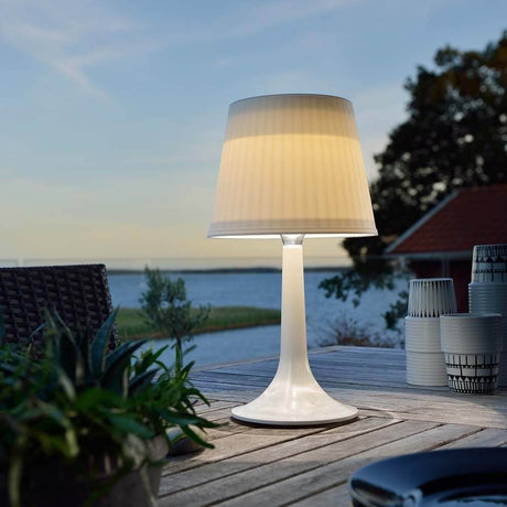 Solar LED table lamp Assisi White on an outdoor table, surrounded by plants and water in the background.