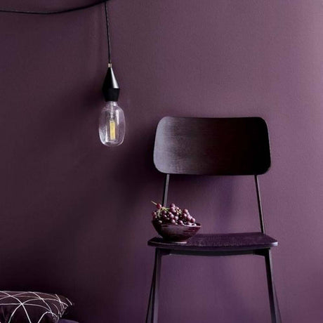 Aud Pendelleuchte Schwarz über einem eleganten schwarzen Stuhl vor lila Wand, ideal für stimmungsvolle Beleuchtung.
