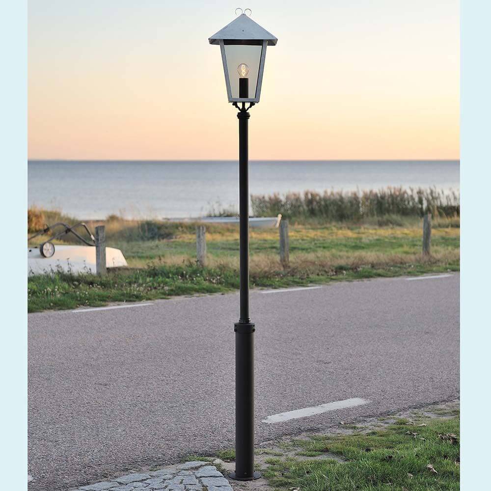 Benu Standleuchte galvanisierter Stahl, rauchfarbenes Acrylglas neben einem Radweg am Meer bei Sonnenuntergang.