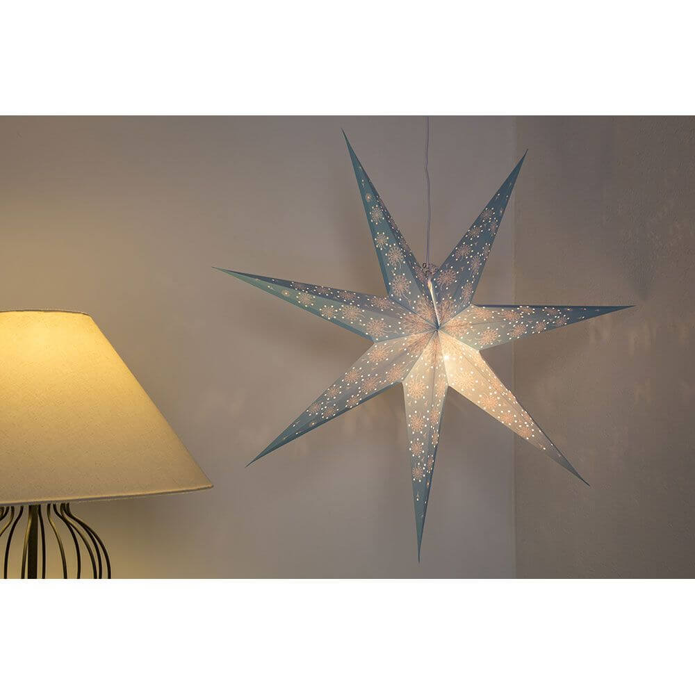 Blauer Papierstern perforiert mit weiß bedrucktem Muster 7 Zacken ohne Leuchmittel an einer Wand, beleuchtet von einer Lampe.