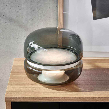 Brokis Tischleuchte Macaron L in rauchigem Glas auf Holzoberfläche, beleuchtet von innen.