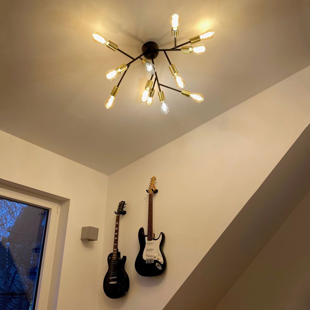 Deckenlampe Heroes 12-flammig Schwarz mit warmem Licht, umgeben von Gitarren an der Wand.