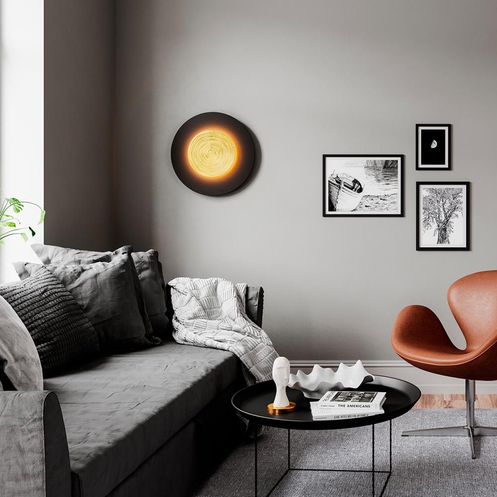 LED-vegglampe Nitra Ø 50 cm svart, gullfarget på grå vegg over en koselig sofa.