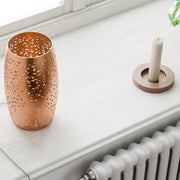 Lámpara de mesa Colby en cobre sobre un alféizar junto a una vela, juego de luz decorativo a través de perforaciones troqueladas.