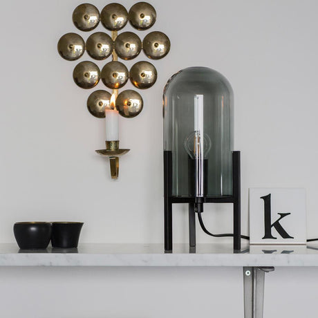 Lampa stołowa Smokey z kloszem szklanym na białej półce, złote kinkiety w tle.