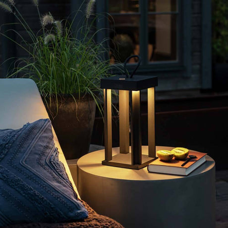 Cannes Outdoor USB lanterne IP54 på et udendørs bord omgivet af planter og en bog.