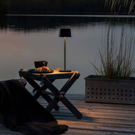 Capri USB-Tischleuchte IP54 auf einem Tisch am Wasser bei Sonnenuntergang, mit dimmbarem Licht.