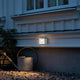 Chieri LED Außen Wandleuchte an einer Hauswand im Freien, in warmem Licht, umgeben von Gartenmaterialien.