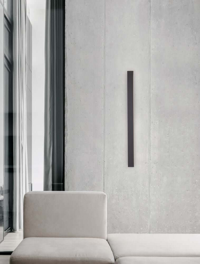 Colby LED Wandleuchte 3000K Schwarz an einer modernen Betonwand neben einem gemütlichen Sofa.