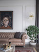 Crown Wandleuchte Gold-Satiniert neben einem braunen Chesterfield-Sofa in einem stilvollen Wohnraum.
