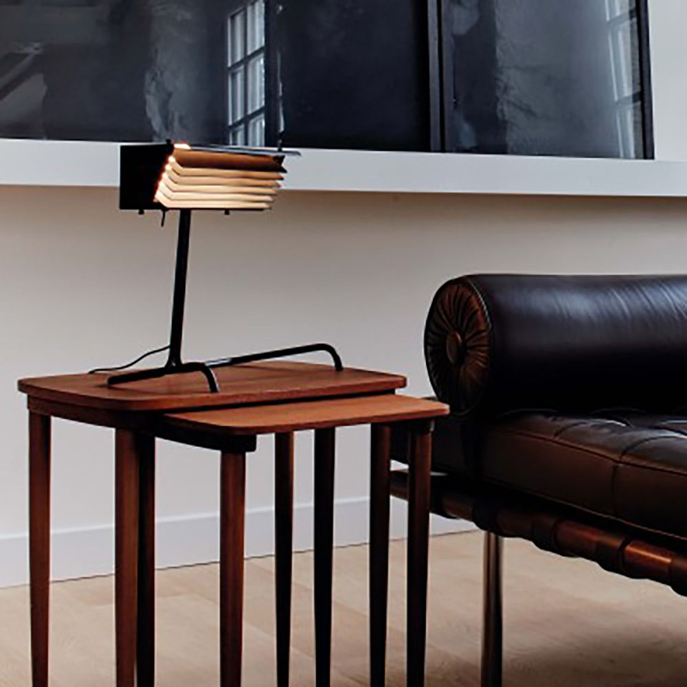 DCWéditions Biny Tischleuchte mit Lamellen auf einem Holztisch neben einem eleganten schwarzen Sofa.