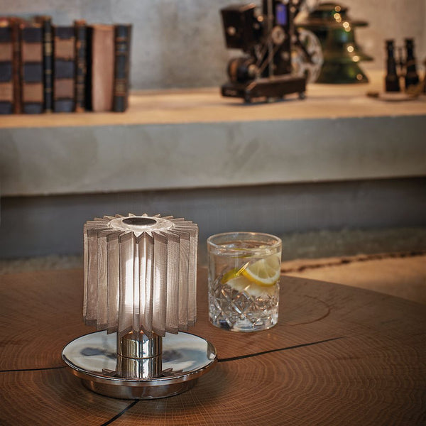 DCWéditions In The Sun Akku LED lampe de table sur une table en bois avec un verre de boisson en arrière-plan.