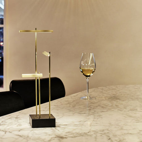 DCWéditions Knokke LED Akku Tischleuchte schwarz Gold auf einem marmorierten Tisch mit einem Weinglas.