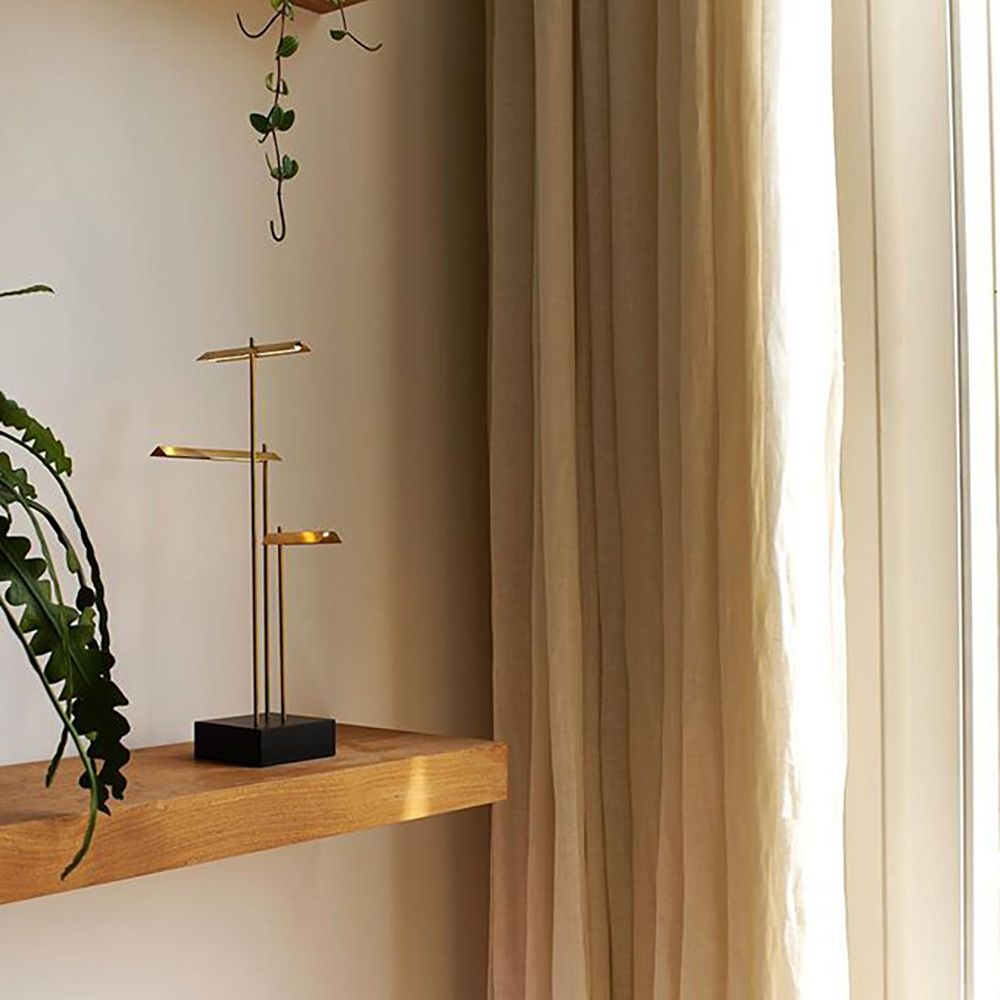 DCWéditions Knokke LED Tischleuchte in schwarz und Gold auf einem Holzregal mit Pflanzen und Vorhängen.