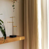 DCWéditions Knokke LED Tischleuchte in schwarz und Gold auf einem Holzregal mit Pflanzen und Vorhängen.