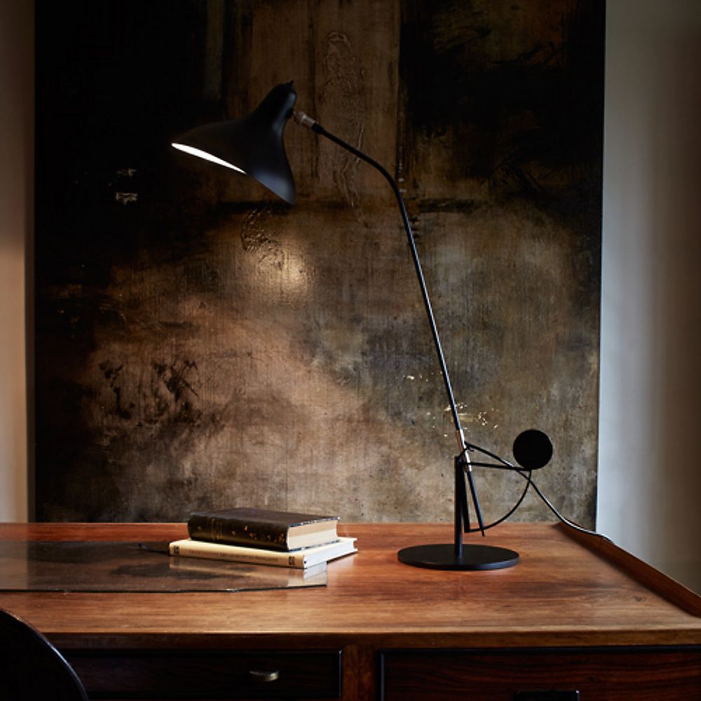 DCWéditions Mantis BS3 Retro Tischleuchte verstellbar auf einem Holztisch mit Büchern und Kunstwerk im Hintergrund.