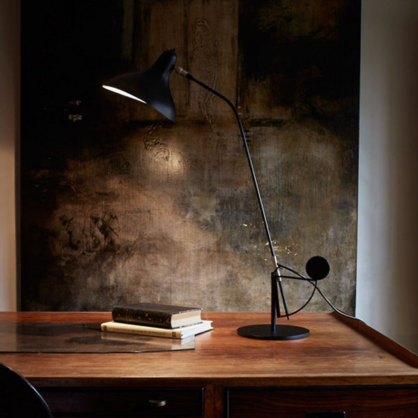 DCWéditions Mantis BS3 Retro Tischleuchte verstellbar auf einem Holzschreibtisch mit Büchern und Kunstwerk im Hintergrund.