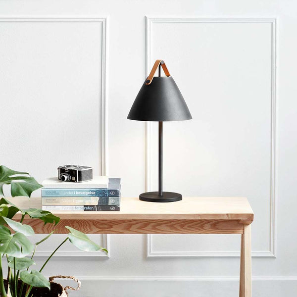 Lampe de table Strap noire sur une table en bois avec des plantes et des livres dans un style nordique moderne.