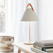 Lampe de table Strap blanche avec poignée en cuir sur une table, design nordique moderne, élégante et fonctionnelle.