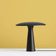 Design Tischlampe Shelter Ø 41cm Schwarz auf weißem Tisch vor gelben Hintergrund.