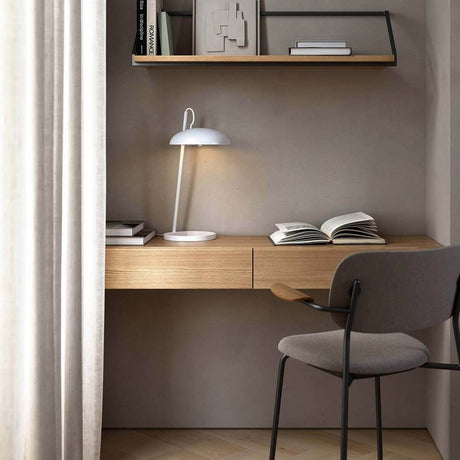 Versale bordlampe på et minimalistisk skrivebord med treoverflate og bokhylle i bakgrunnen.
